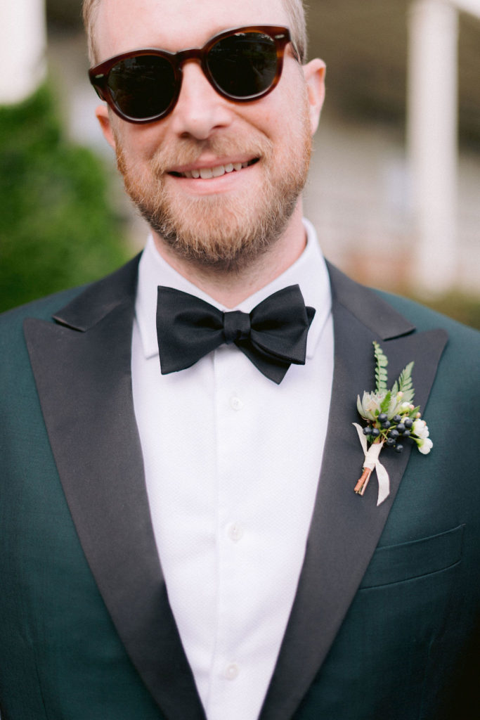 Groom smiles with sunglasses while wearing a green suit and fern boutonniere